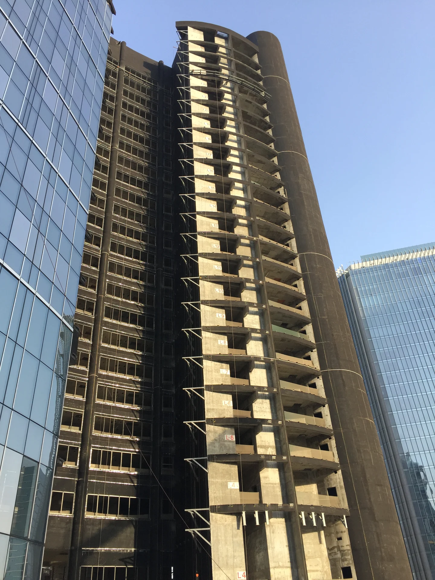 High-rise concrete structure under construction between glazed buildings, with exposed floors and scaffolding.